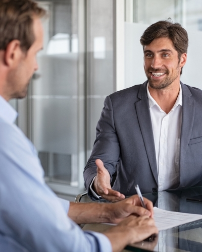 Two men in business attire sit across a desk having a conversation. One man is smiling and gesturing with his hand, while the other holds a pen and a document, suggesting a professional meeting or interview.