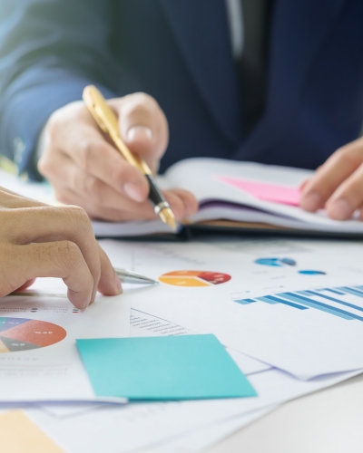 Two people review documents with colorful charts and graphs at a desk; one points with a pen while the other gestures towards the papers, suggesting a business discussion or data analysis.