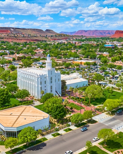Aerial view of the St. George Utah Temple, a white historic building surrounded by green trees and landscaped grounds, set in a city with red rock hills in the background under a partly cloudy sky.