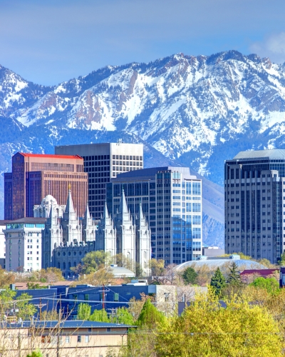 Downtown cityscape with modern and historic buildings, including a white cathedral, set against a backdrop of snow-capped mountains and a partly cloudy sky. Green trees are visible in the foreground.