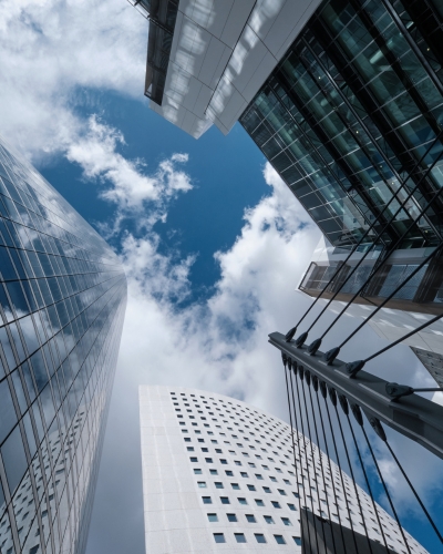 View looking up at modern skyscrapers with glass and concrete facades against a blue sky with scattered clouds; architectural lines create a dramatic, geometric perspective.