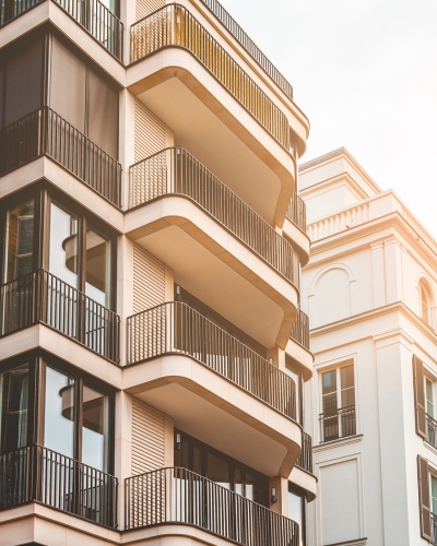 Modern apartment buildings with large balconies and black railings, viewed from a low angle in warm sunlight. The architecture features clean lines and neutral colors, creating a contemporary urban appearance.