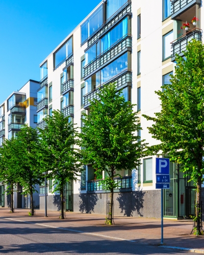 Modern apartment buildings with large windows and balconies line a sunny street. Green trees are planted along the sidewalk, and a blue parking sign is visible in the foreground.
