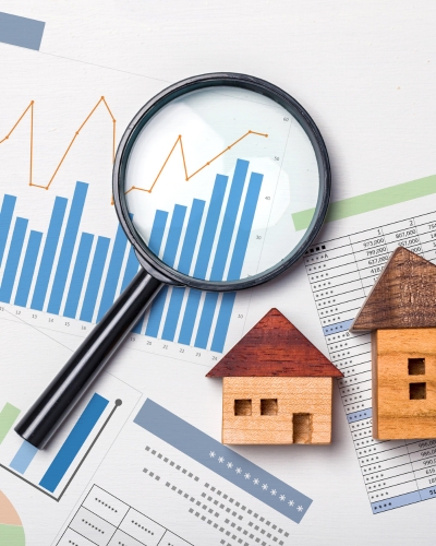 A magnifying glass hovers over bar and line graphs, with two small wooden house models nearby, representing real estate market analysis and data review.
