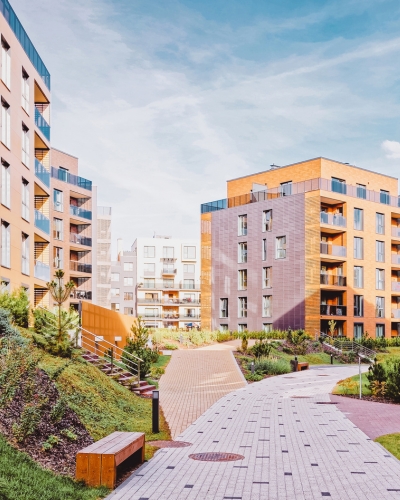 Modern apartment buildings with large windows and balconies surround a landscaped courtyard featuring a paved walkway, greenery, small trees, and wooden benches under a partly cloudy sky.