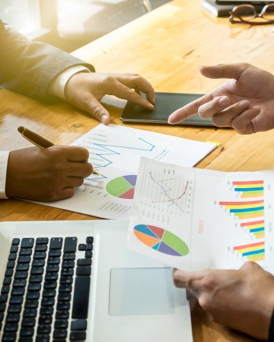 Three people discuss colorful charts and graphs on printed papers at a wooden table, with a laptop, smartphone, and notepad, suggesting a business meeting or data analysis discussion.